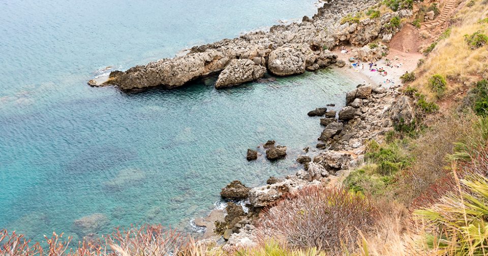 Cala Berretta vista dall'alto. Piccola spiaggia di ghiaia bianca tra due costoni rocciosi, circondata da fitta macchia mediterranea e mare verde smeraldo.