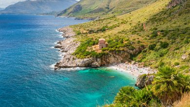 Caletta Tonnarella dell'Uzzo vista dall'alto. Spiaggia di ghiaia bianca circondata da rocce e macchia mediterranea. Un oasi naturale con mare azzurro e verde smeraldo. Sul fondo montagne.