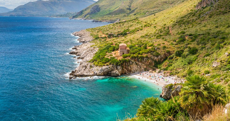 Caletta Tonnarella dell'Uzzo vista dall'alto. Spiaggia di ghiaia bianca circondata da rocce e macchia mediterranea. Un oasi naturale con mare azzurro e verde smeraldo. Sul fondo montagne.