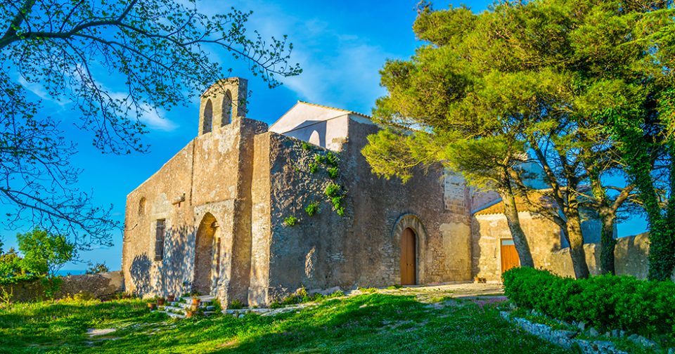 Vista esterna della Chiesa di Sant'Antonio a Erice. Facciate in pietra contornate da alberi e prato verde. Sullo sfondo cielo azzurro.