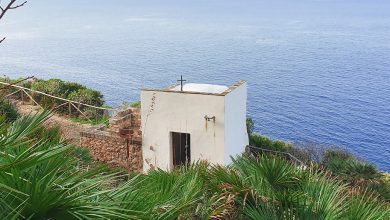La piccola chiesetta bianca del Crocifisso di Cofano vista dall'alto. Tutt'intorno piante verdi di palma nana, sullo sfondo mare e cielo azzurro.