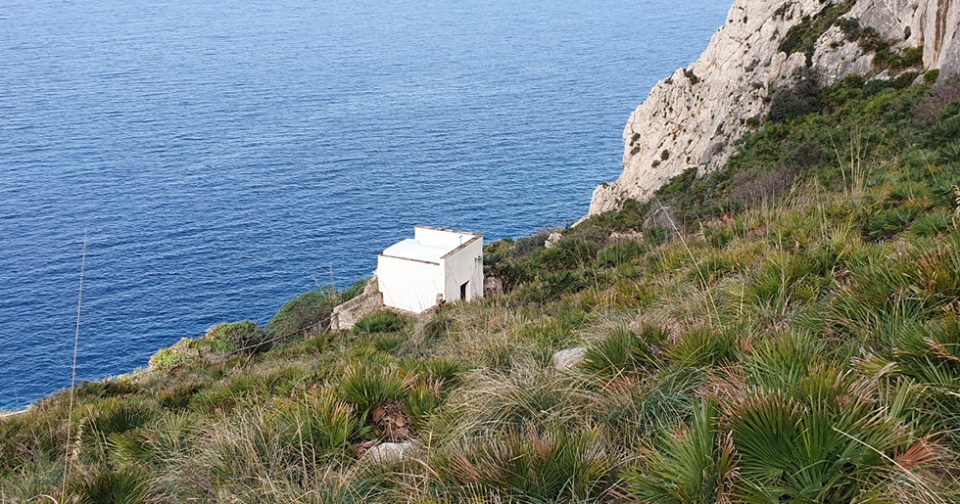 La chiesetta del Crocifisso di Cofano vista dall'alto. Tutt'intono piante di Palma Nana e rocce, sullo sfondo il mare azzurro.