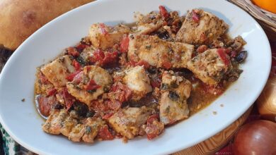 Baccalà alla siciliana con pomodoro, olive e capperi in un piatto ovale bianco, con pane e cipolle sulla tavola