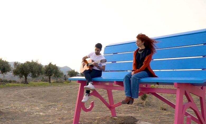 La grande panchina azzurra e fucsia di Centuripe tra gli ulivi, con un ragazzo che suona la chitarra e una ragazza seduta
